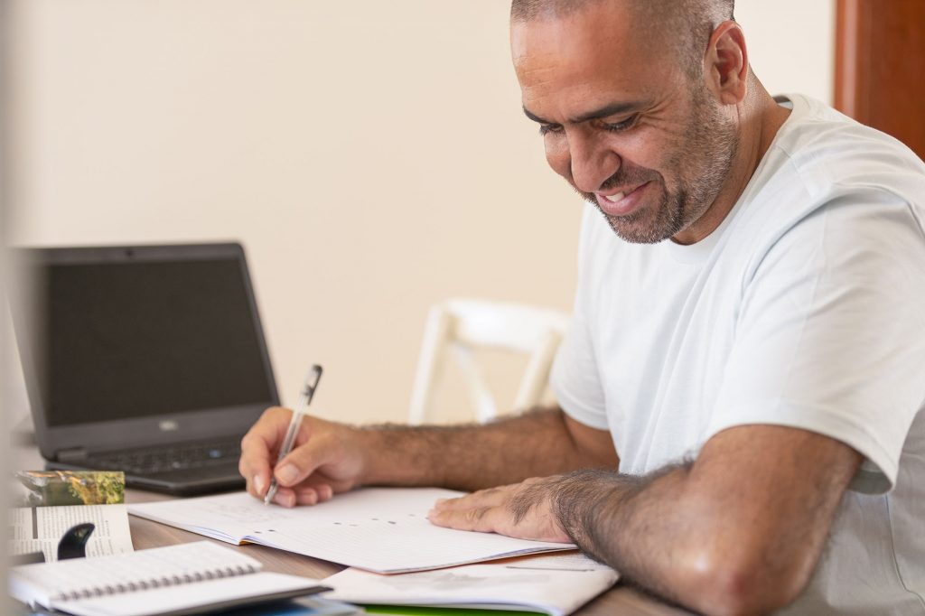 Mature man working at home. He has a laptop and is writing on a document at the table. He could be a businessman at his home office, or a mature age student. He looks happy