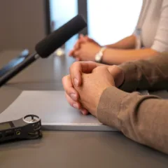 Mãos entrelaçadas sobre uma mesa com um gravador e microfone, em um ambiente de entrevista.