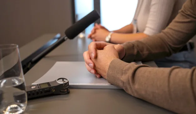 Mãos entrelaçadas sobre uma mesa com um gravador e microfone, em um ambiente de entrevista.