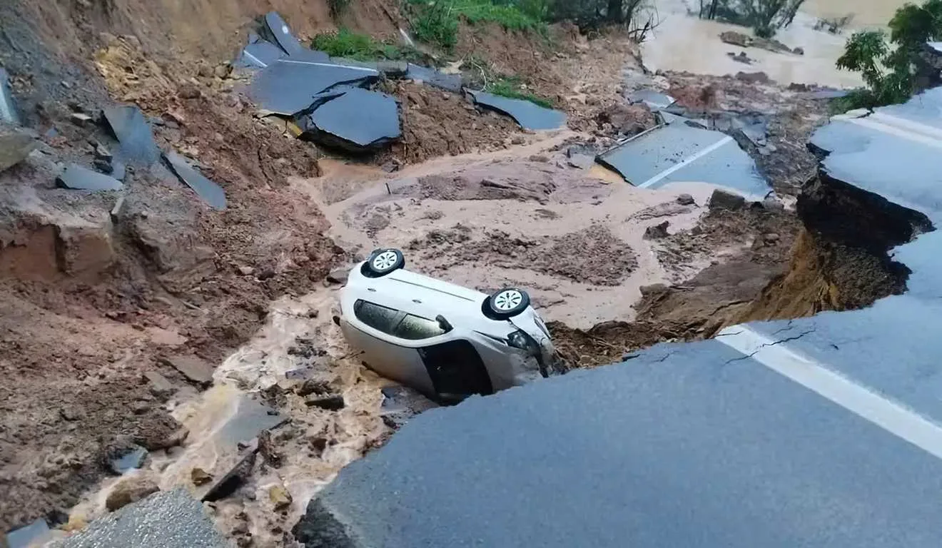 Carro capotado em deslizamento de terra após chuva forte. Estrada destruída e risco iminente.