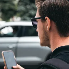Homem usando óculos escuros e segurando um celular perto de carros.