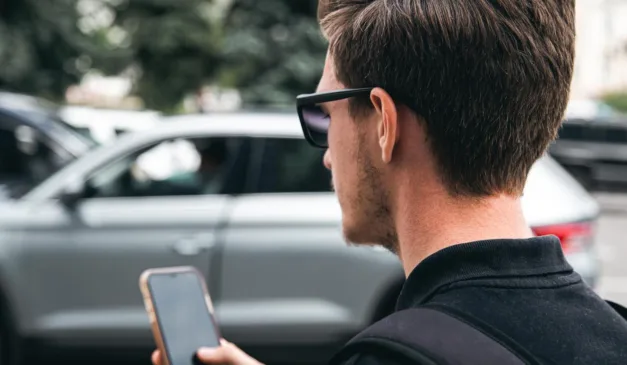 Homem usando óculos escuros e segurando um celular perto de carros.