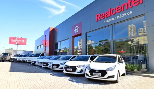 Fachada da Realcenter com carros brancos estacionados em frente, sob um céu azul.