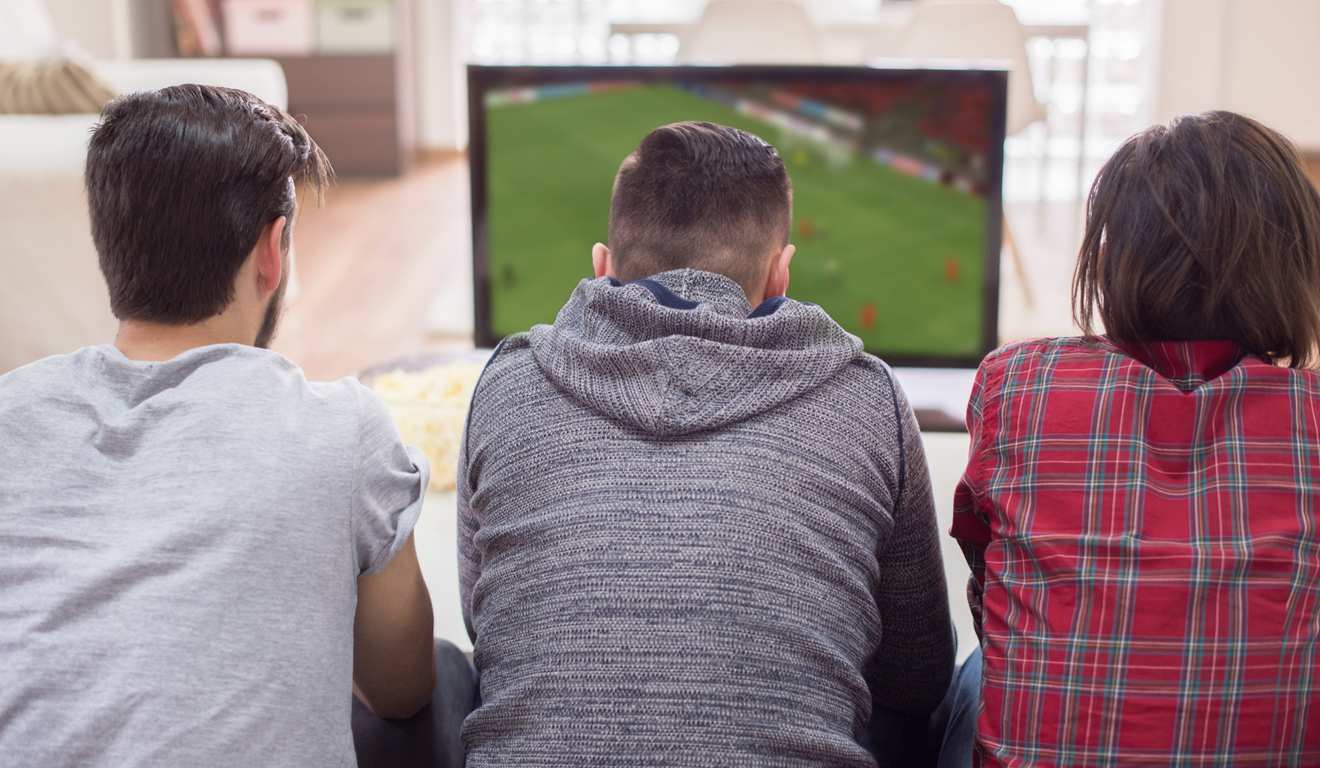 Três pessoas assistindo TV juntas. Uma partida de futebol é exibida na tela.