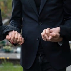 Três pessoas de luto de mãos dadas em um cemitério, usando roupas pretas.