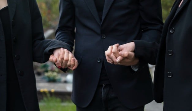 Três pessoas de luto de mãos dadas em um cemitério, usando roupas pretas.