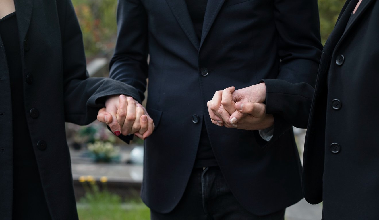 Três pessoas de luto de mãos dadas em um cemitério, usando roupas pretas.