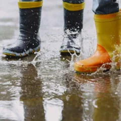 Crianças brincando na chuva com botas coloridas, respingando água.