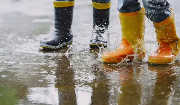 Crianças brincando na chuva com botas coloridas, respingando água.