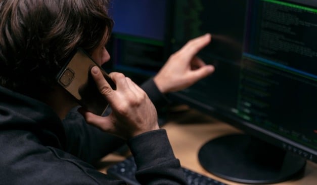 Homem falando ao telefone enquanto trabalha em um computador com código na tela.