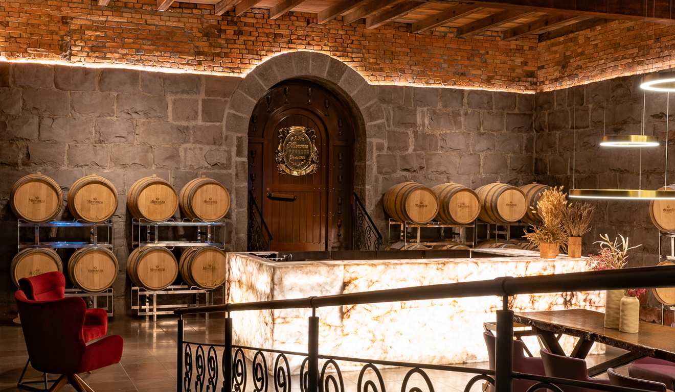 Interior de adega com barris de vinho, porta de madeira e iluminação ambiente.