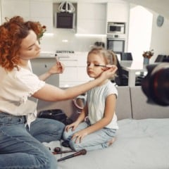 Mãe aplica maquiagem em sua filha em um ambiente doméstico, com uma câmera em foco.