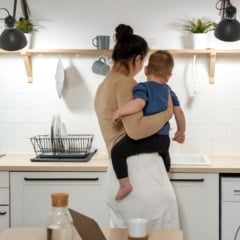 Mãe segura criança na cozinha moderna com armários brancos e eletrodomésticos.