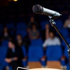 Microfone em palco de auditório com pessoas sentadas em cadeiras azuis no fundo, desfocadas.