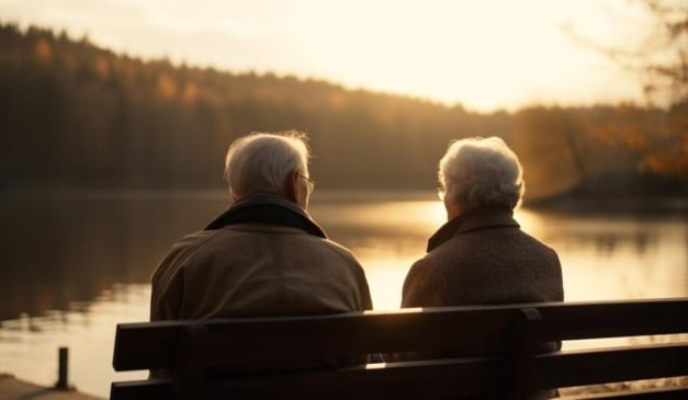 Casal de idosos sentado em um banco à beira do lago, apreciando o pôr do sol.