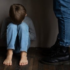 Criança encolhida no chão com a cabeça entre os joelhos e pessoa em pé.