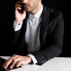 Homem de terno usando laptop e falando ao telefone em fundo preto.