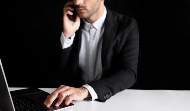 Homem de terno usando laptop e falando ao telefone em fundo preto.