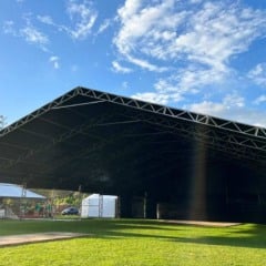 Tenda grande com estrutura metálica e cobertura escura em um gramado verde sob um céu azul com nuvens.