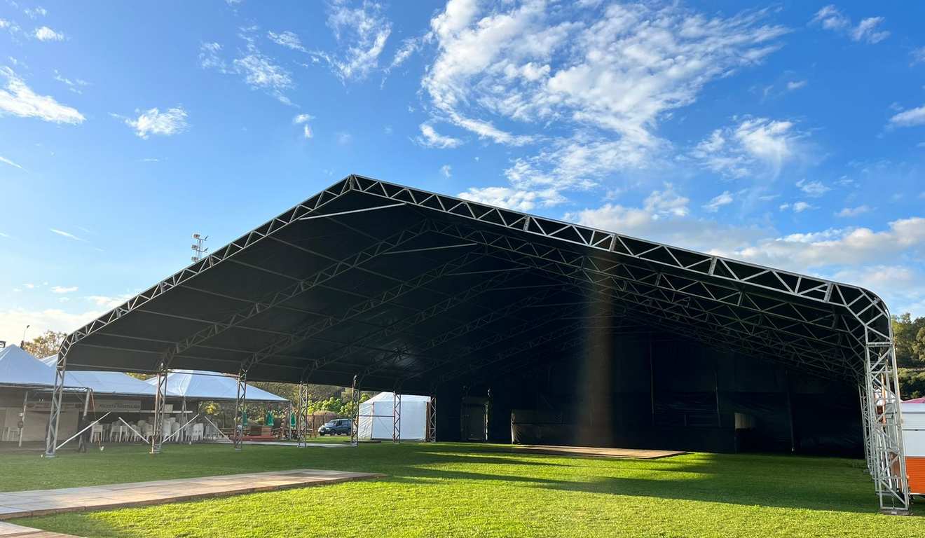 Tenda grande com estrutura metálica e cobertura escura em um gramado verde sob um céu azul com nuvens.