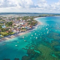 Vista aérea de uma cidade costeira com praia, mar azul e barcos à vela.