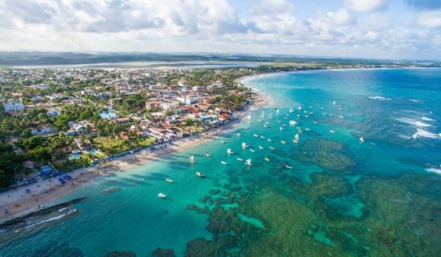 Vista aérea de uma cidade costeira com praia, mar azul e barcos à vela.