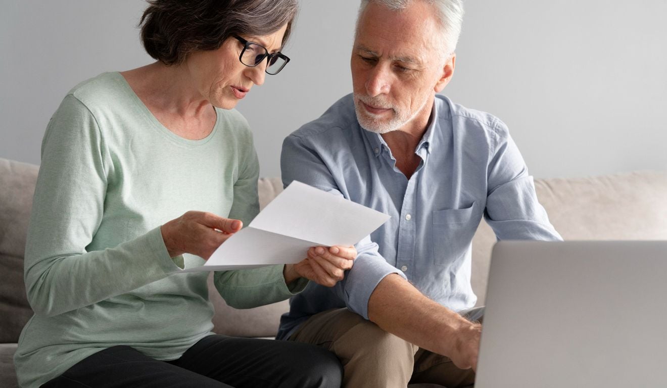 Casal de idosos analisando documentos e usando um laptop em casa.