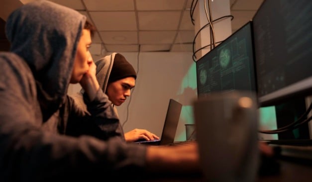 Dois jovens com capuzes trabalhando em computadores, possivelmente hackers ou programadores.
