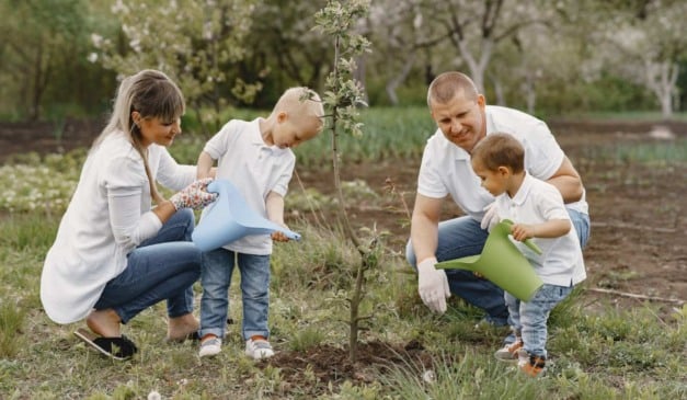 Família plantando árvore, pai, mãe e filhos regando a muda juntos no jardim.