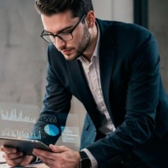 Homem de terno analisa gráficos em tablet, com óculos e iluminação de escritório.