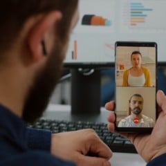 Homem em videochamada com duas pessoas no smartphone. Monitor e teclado ao fundo.