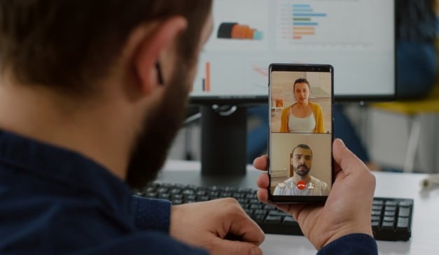 Homem em videochamada com duas pessoas no smartphone. Monitor e teclado ao fundo.