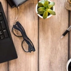 Laptop, óculos, caneta, bloco de notas, xícara de café e planta em mesa de madeira.