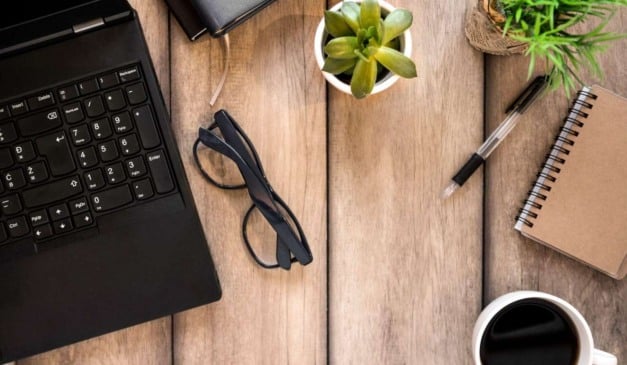 Laptop, óculos, caneta, bloco de notas, xícara de café e planta em mesa de madeira.