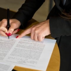 Mãos femininas assinando um documento com unhas vermelhas e uma caneta preta em um ambiente de escritório.