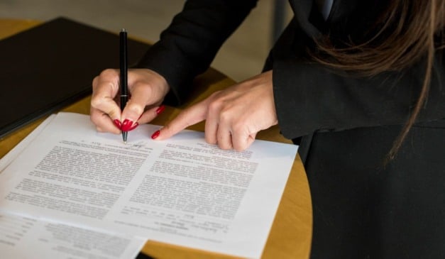 Mãos femininas assinando um documento com unhas vermelhas e uma caneta preta em um ambiente de escritório.