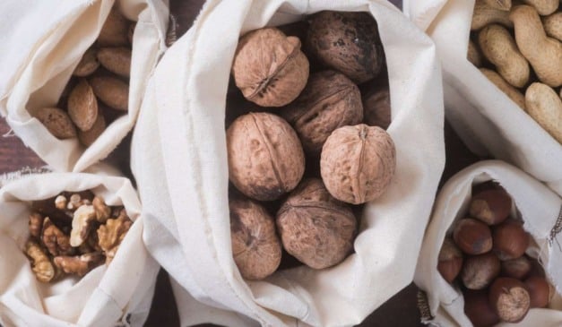 Sacos de tecido repletos de nozes, amêndoas, amendoins, avelãs e nozes descascadas, dispostos em uma superfície escura.