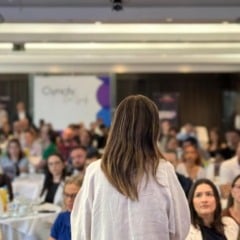 Palestrante fala para plateia em evento corporativo, vista por tr&aacute;s, com foco na audi&ecirc;ncia.