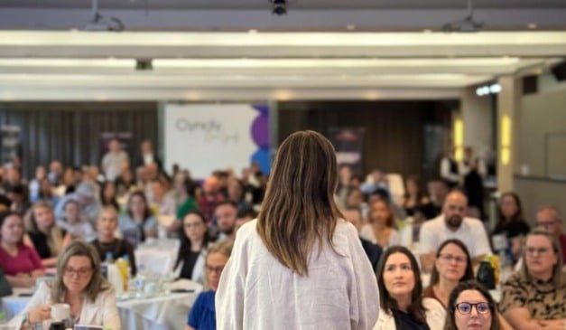 Palestrante fala para plateia em evento corporativo, vista por trás, com foco na audiência.