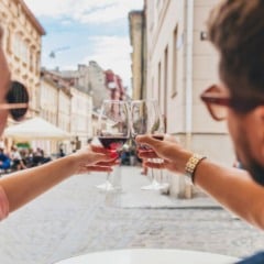 Casal brindando com vinho em um restaurante ao ar livre, com edif&iacute;cios hist&oacute;ricos ao fundo.
