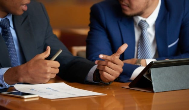 Dois homens de terno discutindo em uma reunião de negócios, com documentos e tablet na mesa.