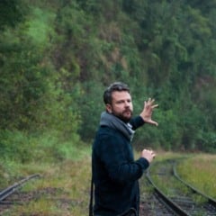 Homem de barba aponta para a natureza ao lado de trilhos de trem em ambiente verde.