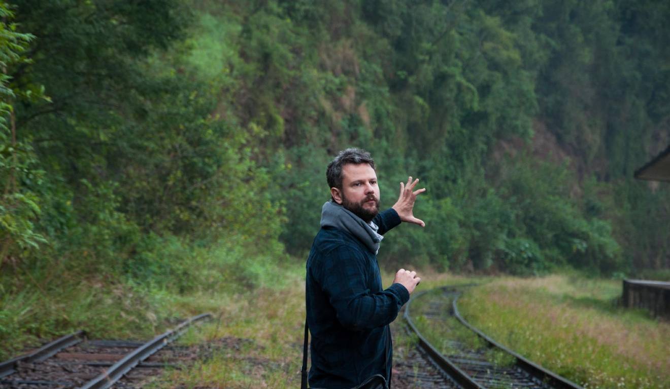 Homem de barba aponta para a natureza ao lado de trilhos de trem em ambiente verde. Homem de barba aponta para a natureza ao lado de trilhos de trem em ambiente verde.