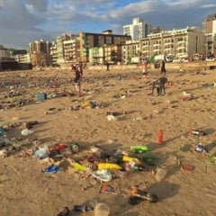 Praia coberta por lixo ap&oacute;s evento, com pr&eacute;dios ao fundo.