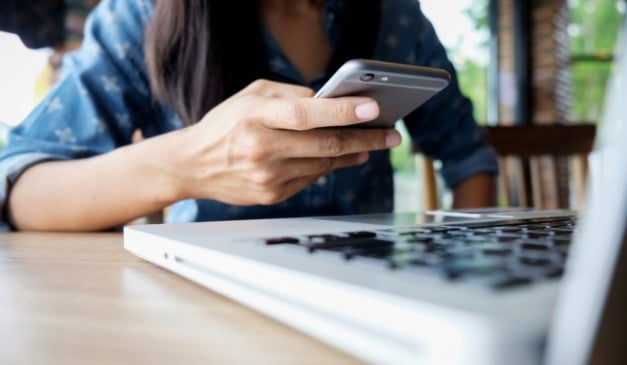 Uma pessoa usando um smartphone em cima de uma mesa com um laptop.