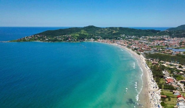 Vista aérea da praia com mar azul turquesa, areia e cidade ao fundo.