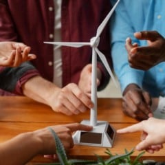 Grupo de pessoas reunidas em torno de uma maquete de turbina e&oacute;lica, com dedos apontando.