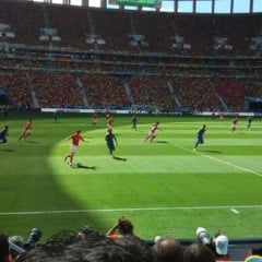 Jogo de futebol em est&aacute;dio lotado. Jogadores correndo em campo verde.